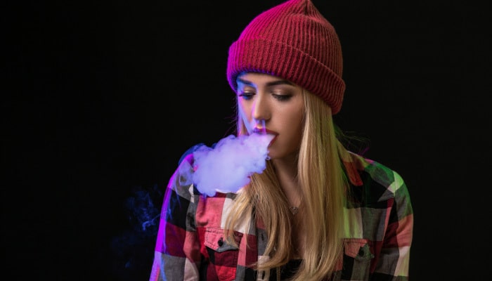 Woman with red beanie and flannel shirt against black background blowing out vapor from a Boston THC Vape Cart.