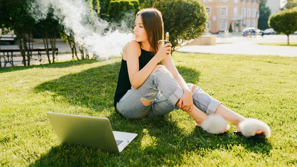 A woman sitting outside on the grass with her laptop enjoying the Lancaster THCa vape cart.
